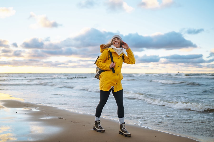 Frau in gelber Jacke geht am Strand entlang und lächelt, im Hintergrund Meer und Wolkenhimmel.